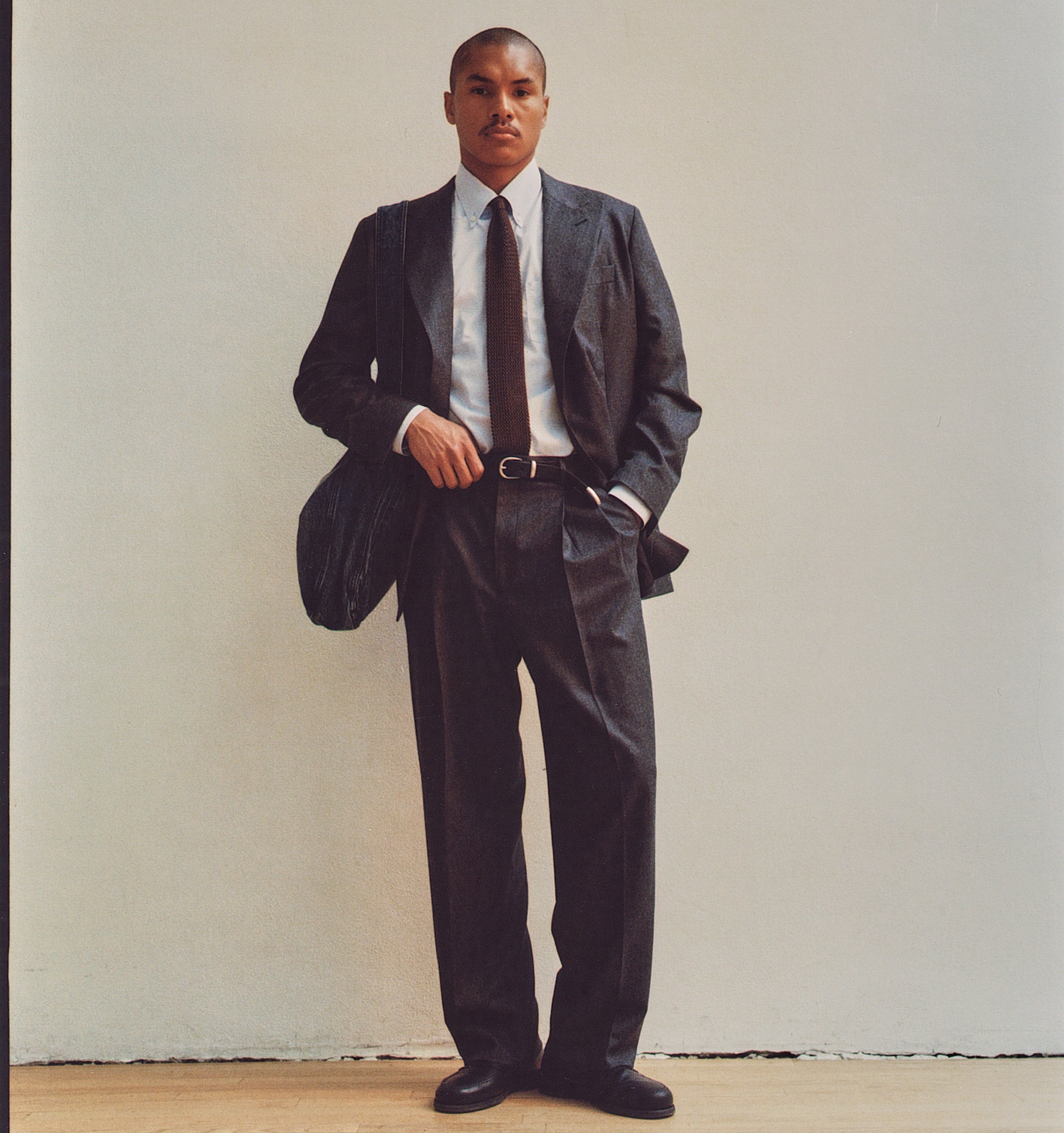 A man stands against a plain off-white wall wearing a dark charcoal gray suit, a white dress shirt, a dark brown tie, and black dress shoes. He is holding a black shoulder bag.